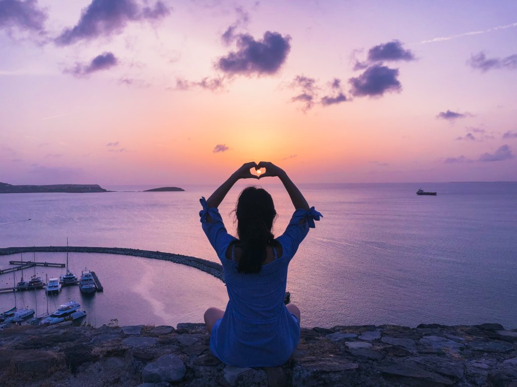 lady sitting at beach with a pink purple sunset holding her hands above her in a heart shape for chakra mindset